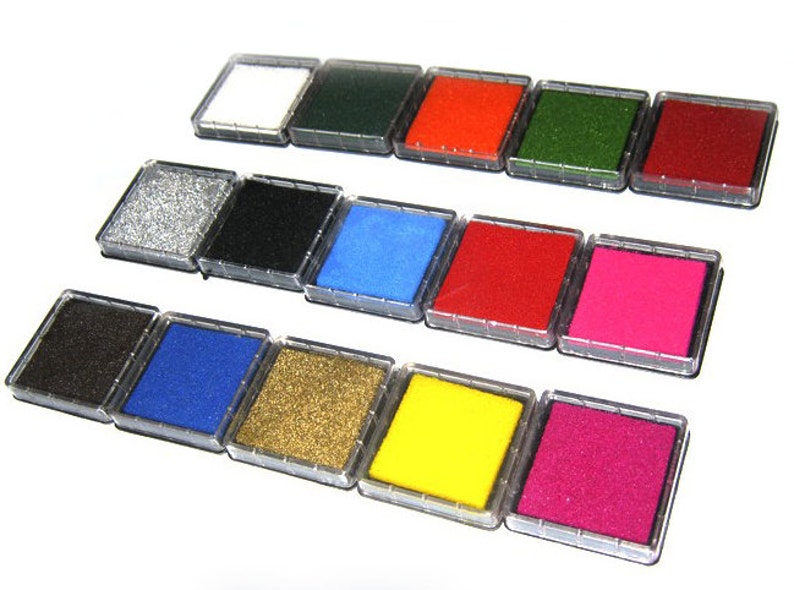 May include: A collection of ink pads in various colors, including white, green, red, and pink. Each pad is square and housed in a clear plastic case. The ink pads are arranged in rows, showcasing a range of vibrant hues suitable for crafts and stamping.