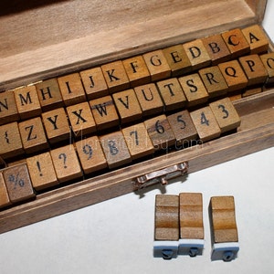 May include: A wooden box filled with letter and number stamps. The stamps are made of wood and have black lettering. The box is open and the stamps are visible. There are three additional stamps outside of the box.