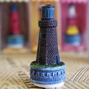 Puede incluir: Una figurita de faro en miniatura con un exterior de ladrillo, un techo azul y una base con un diseño de piedra. El faro está sobre una superficie marrón.