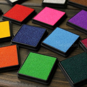 Könnte beinhalten: Eine Sammlung von zehn Stempelkissen in verschiedenen Farben, darunter Rot, Pink, Blau, Grün, Orange, Gelb, Lila und Dunkelgrün. Die Stempelkissen sind quadratisch und haben einen schwarzen Kunststoffrahmen.