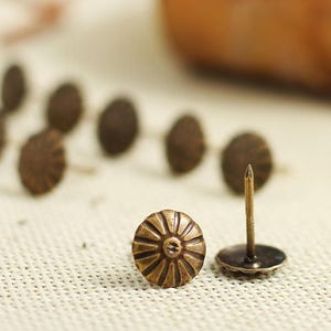 Puede incluir: Primer plano de una sola tachuela de lat&oacute;n antiguo con una cabeza circular decorativa. La tachuela est&aacute; sobre una superficie de tela blanca, rodeada de otras tachuelas similares.