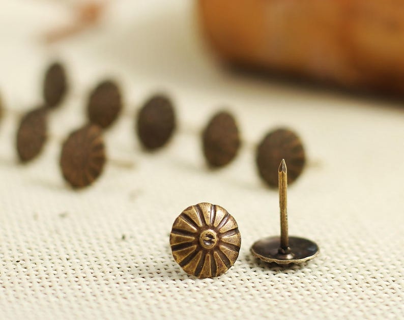 Puede incluir: Primer plano de una sola tachuela de lat&oacute;n antiguo con una cabeza circular decorativa. La tachuela est&aacute; sobre una superficie de tela blanca, rodeada de otras tachuelas similares.