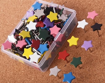 May include: A selection of star-shaped push pins in a clear plastic container and scattered on a brown corkboard. The pins are in various colours, including yellow, pink, blue, and white.