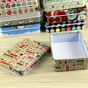 May include: A set of small, rectangular tin boxes with various colorful patterns. The boxes are stacked on top of each other. The top box is open and empty, revealing a white interior. The boxes feature different designs, including polka dots, stripes, and numbers.
