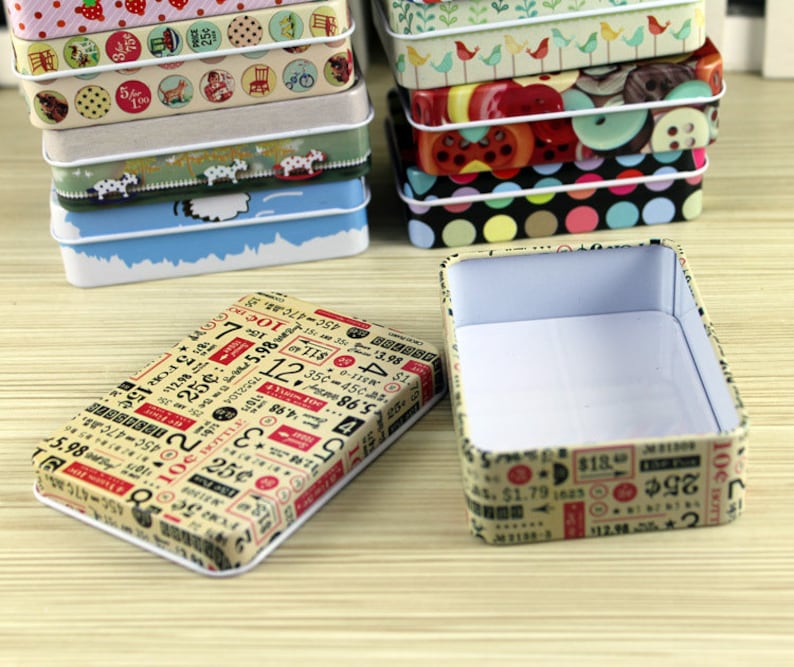 May include: A set of small, rectangular tin boxes with various colorful patterns. The boxes are stacked on top of each other. The top box is open and empty, revealing a white interior. The boxes feature different designs, including polka dots, stripes, and numbers.