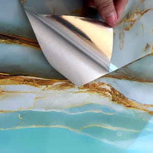 Peut inclure: Gros plan sur un papier contact marbr&eacute; turquoise et or avec un dos argent&eacute;. Le dos est d&eacute;coll&eacute; pour r&eacute;v&eacute;ler le c&ocirc;t&eacute; adh&eacute;sif du papier.