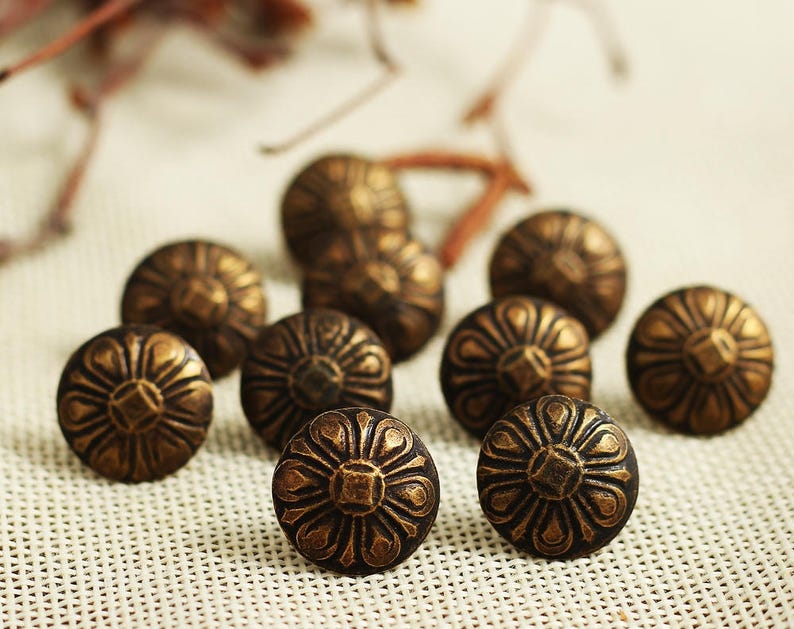 Puede incluir: Un grupo de diez tachuelas decorativas de metal de color dorado antiguo con un dise&ntilde;o floral. Las tachuelas tienen un centro elevado y una parte posterior plana.