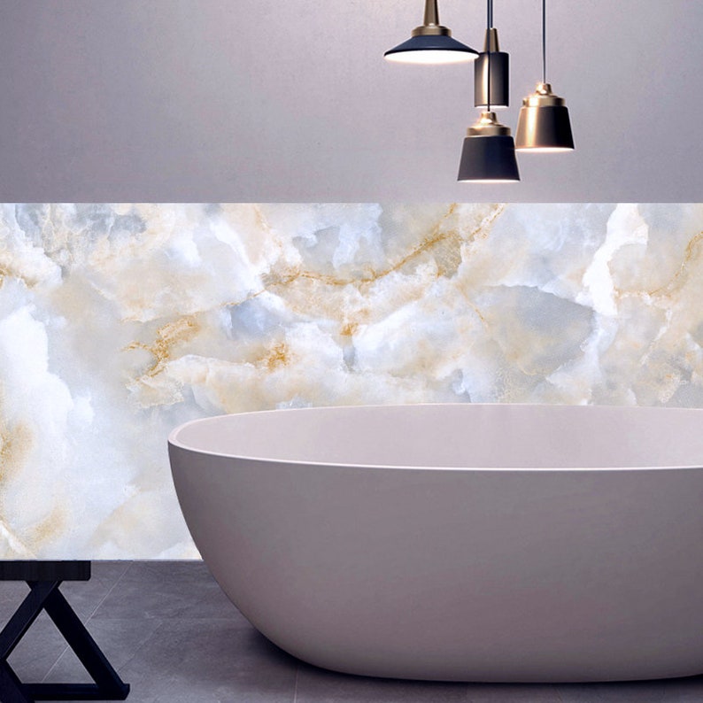 May include: A white freestanding bathtub with a light blue and gold marble wall behind it. Three black and gold pendant lights hang from the ceiling above the bathtub.