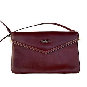 May include: A deep burgundy envelope-style clutch bag with a smooth finish, featuring a thin wrist strap and a long shoulder strap. The front flap has a V-shaped edge with gold-tone metal accents and a central oval logo plaque with gold lettering.