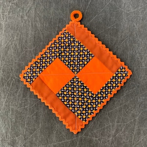 Puede incluir: Un salvamanteles cuadrado y acolchado con un diseño de rombos. El diseño presenta cuadrados naranjas y tela negra con un estampado de lunares amarillos y blancos. Los bordes son festoneados y naranjas, con un pequeño lazo naranja para colgar.