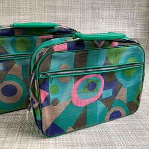 May include: Two rectangular travel cases featuring a geometric pattern in shades of teal, green, brown, and pink. Each case has a bright green plastic handle, a zippered main compartment, and a smaller zippered front pocket with green trim.