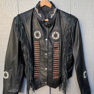 Könnte beinhalten: Schwarze Lederjacke mit Fransen und dekorativen Perlen. Die Jacke hat eine Knopfleiste, einen Kragen mit farbenfrohen Perlen und dekorative kreisförmige und vertikale Muster. Die Ärmel und Schultern haben Fransen.
