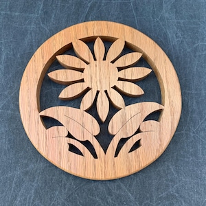 May include: A circular wooden trivet featuring a cutout design of a flower with twelve petals and two large leaves at the base. The natural light brown wood grain is visible throughout the piece, which is framed by a solid circular border.