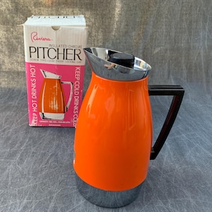 May include: A bright orange insulated pitcher with a chrome base, a chrome rim, and a black handle. The pitcher includes a black screw-type lid. Beside it stands the original white and pink box labeled Riviera Insulated Chrome Pitcher.