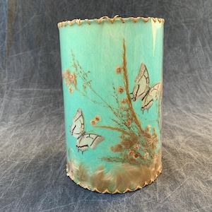 May include: A cylindrical lampshade featuring a vibrant turquoise background decorated with two detailed butterflies and brown dried floral sprigs. The top and bottom edges are finished with a scalloped trim in a light tan color.