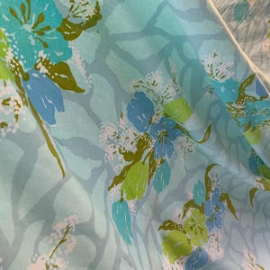 Peut inclure: Gros plan sur un tissu à motifs floraux. Le tissu présente de grandes fleurs turquoise et bleues avec des feuilles vertes et des accents blancs sur un fond bleu clair. Le tissu est plié, montrant sa texture.