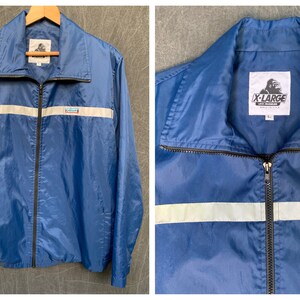 May include: A blue nylon jacket with a black zip and a white reflective stripe across the chest. The jacket has a logo on the left chest that says "X-Large" and a logo on the right chest that says "K-Mart".