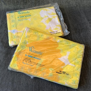 Puede incluir: Dos paquetes de sábanas planas gemelas Wamsutta Ultracale. Las sábanas están en envases de plástico transparente. Las sábanas son amarillas con un diseño floral naranja y blanco. El embalaje tiene el texto "NO IRONING NECESSARY".