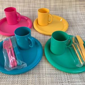 May include: A set of four plastic dinnerware place settings in pink, yellow, teal, and green. Each setting includes a round plate, a matching mug, and a clear plastic bag containing a fork and a knife. The items are arranged in a two-by-two grid.