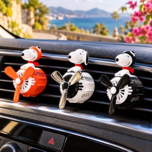 May include: Three Snoopy car air fresheners attached to a vehicle air vent. Each features a white Snoopy figure wearing a pilot cap and red scarf, mounted on a spherical base with a rotating propeller. The bases are orange, white, and black.