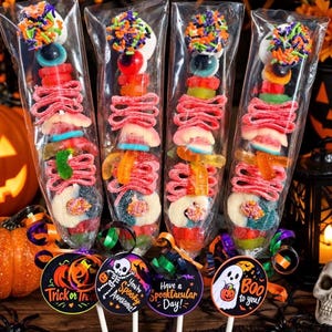 May include: Four Halloween candy skewers in clear plastic wrappers, each topped with a marshmallow and colorful sprinkles. The skewers feature layers of gummy worms, sour belts, and fruit-flavored candies, with round tags attached to the sticks.