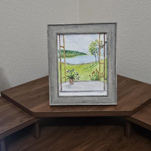 Könnte beinhalten: Ein gerahmtes Aquarellbild, das den Blick aus einem offenen Fenster auf eine grüne Landschaft mit einem See, Bäumen und einer Topfpflanze mit violetten Blüten auf dem Sims zeigt. Der Rahmen besteht aus verwittertem grauem Holz. Signatur unten rechts.