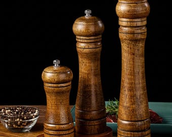 May include: Three wooden pepper mills of varying heights stand on a wooden base. Each mill features a dark brown wood grain finish, a rounded top, and a small silver-coloured metal knob at the very peak for adjusting the grinding mechanism.