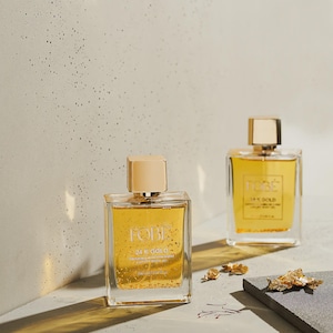 Può includere: Due flaconi di profumo quadrati in vetro con tappi dorati contenenti liquido ambrato e scaglie d'oro. Le etichette riportano la scritta FOBE 24 K GOLD SAFFRON & MANDARIN ZEST LUXURY BODY OIL. Piccoli pezzi di foglia d'oro sono sparsi vicino.