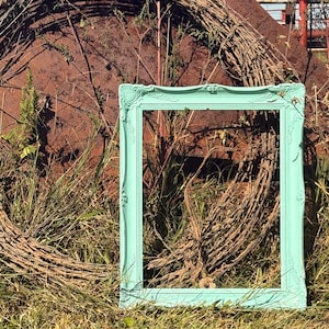 May include: A rectangular picture frame painted in a bright mint green color. The frame features ornate, raised decorative carvings along its edges and corners, showcasing a smooth finish that contrasts with the surrounding natural environment.
