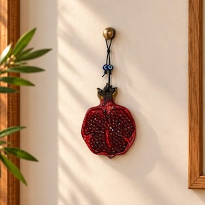May include: A wall-hanging ornament shaped like a halved pomegranate with a glossy red finish and visible seeds. It hangs from a black cord featuring two small blue and white evil eye beads, attached to a circular brass-colored wall hook.