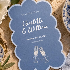 Pode incluir: Um cartão Save the Date azul com bordas recortadas, apresentando texto em branco que diz Save the Date, Charlotte & William, Saturday, July 17, 2027, Tuscany, Italy, e uma ilustração linear de duas taças de champanhe.