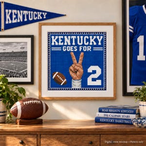 May include: A square framed cross-stitch pattern featuring a blue background with the text KENTUCKY GOES FOR 2. The design includes a hand making a peace sign, a brown football, and the number 2 in white, all rendered in a pixelated style.