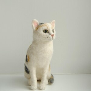 May include: A needle-felted cat figurine featuring white fur with patches of orange and dark gray. The cat has green eyes, a pink nose, and long white whiskers, sitting in an upright position with its head turned slightly to the right side.