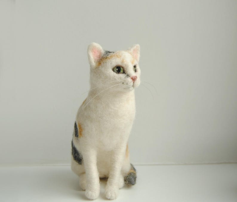 May include: A needle-felted cat figurine featuring white fur with patches of orange and dark gray. The cat has green eyes, a pink nose, and long white whiskers, sitting in an upright position with its head turned slightly to the right side.