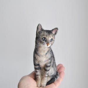 May include: A needle-felted gray tabby cat figurine with dark gray stripes, a light cream-colored chest, pointed ears, and blue eyes. The cat is depicted in a seated position with a small pink nose and long white whiskers extending from its muzzle.