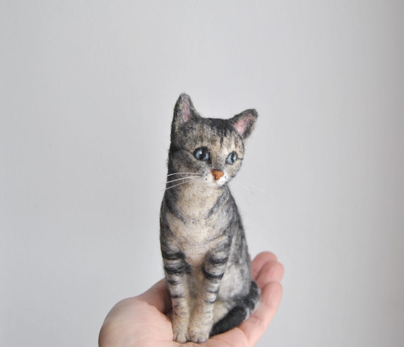 May include: A needle-felted gray tabby cat figurine with dark gray stripes, a light cream-colored chest, pointed ears, and blue eyes. The cat is depicted in a seated position with a small pink nose and long white whiskers extending from its muzzle.