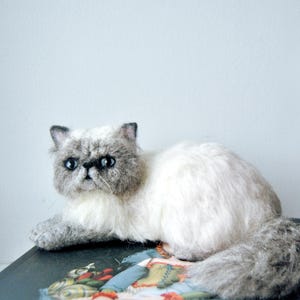 May include: A needle-felted cat figurine with a cream-colored body and a grey face, ears, and tail. The cat has dark, round eyes, a small black nose, and a textured, fluffy appearance. It is posed in a lying position with its tail curled.