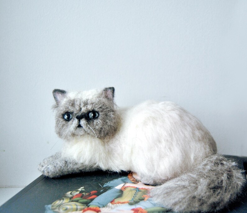 May include: A needle-felted cat figurine with a cream-colored body and a grey face, ears, and tail. The cat has dark, round eyes, a small black nose, and a textured, fluffy appearance. It is posed in a lying position with its tail curled.