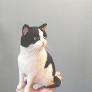 May include: A needle felted cat figurine featuring a black and white coat with a white blaze on its face, pointed ears with pink inner details, dark eyes, and long thin white whiskers. The cat is depicted in a seated position with its tail curled.