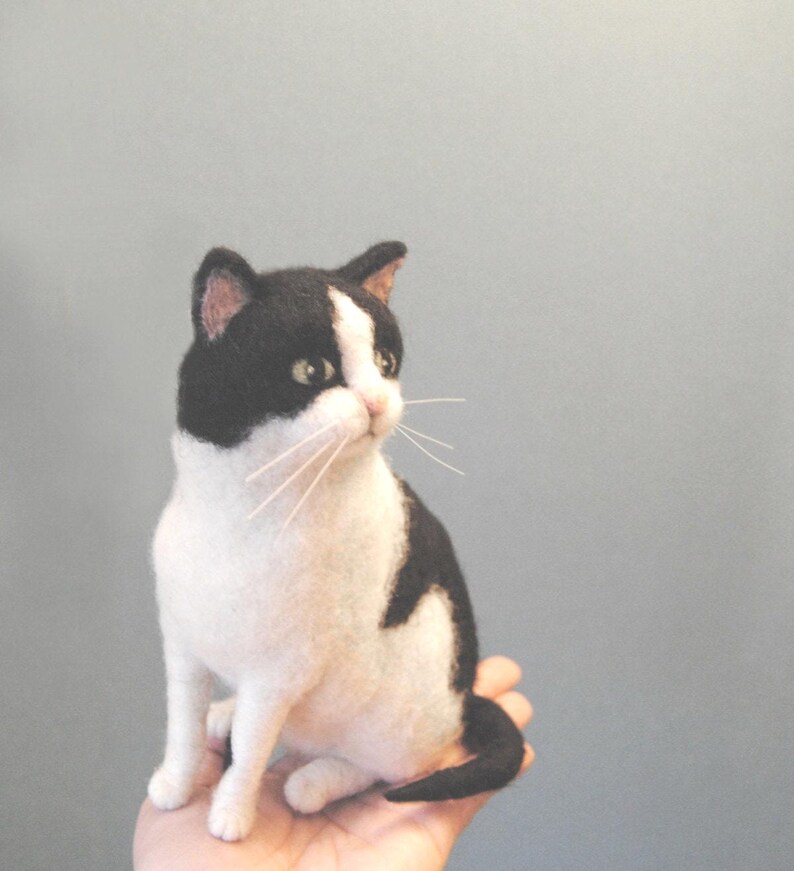 May include: A needle felted cat figurine featuring a black and white coat with a white blaze on its face, pointed ears with pink inner details, dark eyes, and long thin white whiskers. The cat is depicted in a seated position with its tail curled.