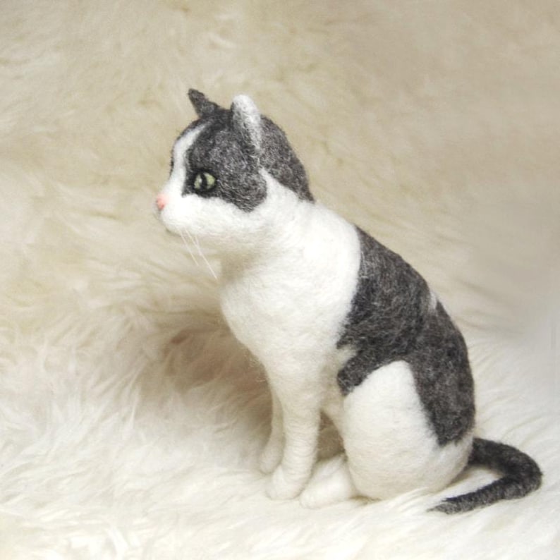 May include: A needle felted cat figurine featuring a white chest, face, and legs with dark gray patches on the head, back, and tail. The cat has a small pink nose, green eyes, and long white whiskers, captured in a seated position with a textured finish.
