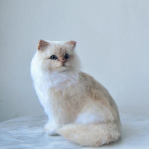 May include: A needle felted cat figurine with white and light tan fur. The cat has dark eyes, a small pinkish nose, and pointed ears. It is depicted in a seated position with its tail curled around its body, showcasing a soft, textured appearance.