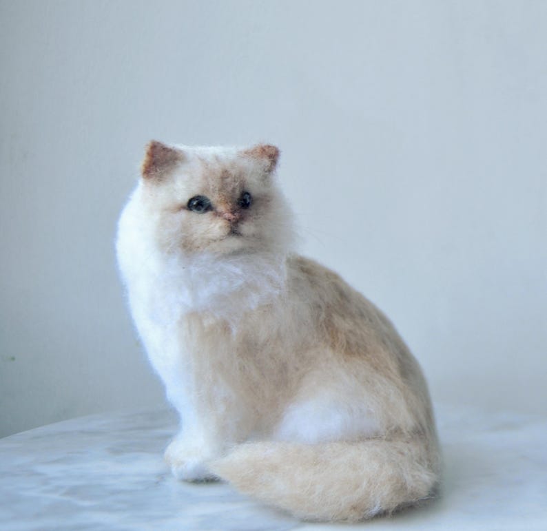 May include: A needle felted cat figurine with white and light tan fur. The cat has dark eyes, a small pinkish nose, and pointed ears. It is depicted in a seated position with its tail curled around its body, showcasing a soft, textured appearance.