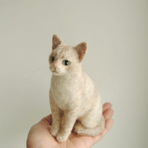 May include: A needle felted cat figurine with light beige and cream fur, pointed ears, and green eyes. The small sculpture features a realistic texture, a pink nose, and thin whiskers extending from the muzzle, captured in a seated position.