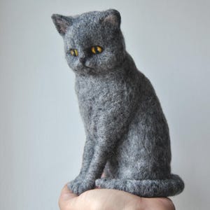 May include: A needle-felted sculpture of a grey cat with a textured wool finish. The cat is depicted in a seated position with its tail curled around its base. It features prominent, round, bright yellow eyes and small, pointed ears.