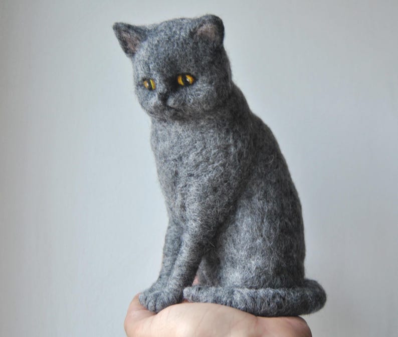 May include: A needle-felted sculpture of a grey cat with a textured wool finish. The cat is depicted in a seated position with its tail curled around its base. It features prominent, round, bright yellow eyes and small, pointed ears.