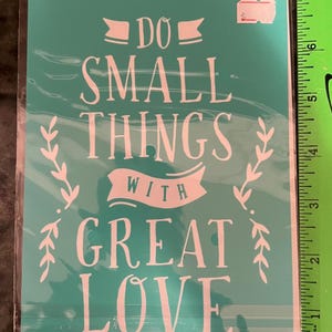 May include: A teal rectangular stencil in a clear plastic sleeve featuring the white text DO SMALL THINGS WITH GREAT LOVE and two leafy vine illustrations. A green ruler on the right shows the stencil is approximately 8.5 inches tall.