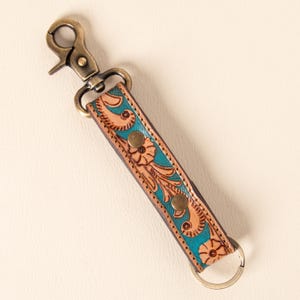 May include: A leather keychain featuring a tan strap with an embossed floral pattern and a vibrant turquoise background. The strap is secured with two brass-colored metal rivets and includes a matching brass-colored swivel lobster clasp and ring.