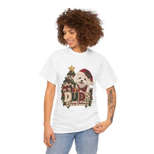 May include: A white short-sleeve t-shirt featuring a graphic of a white dog wearing a red plaid Santa hat and scarf, positioned in front of a Christmas tree and gifts, with the text DUDE and Merry Christmas printed below.