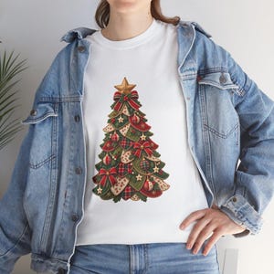 May include: A white t-shirt featuring a graphic of a Christmas tree decorated with red bows, patterned fabric patches, and a gold star topper. The shirt is worn under a blue denim jacket with metal buttons and visible stitching details.